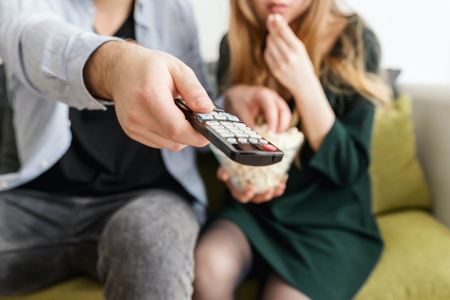 Close up photo of a couple on the couch, the man holding a remote control and pointing it at the camera.
